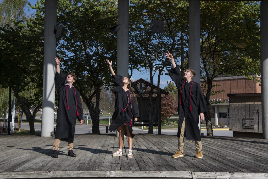 Three graduates wearing regalia and red cords toss their graduation cap in the air