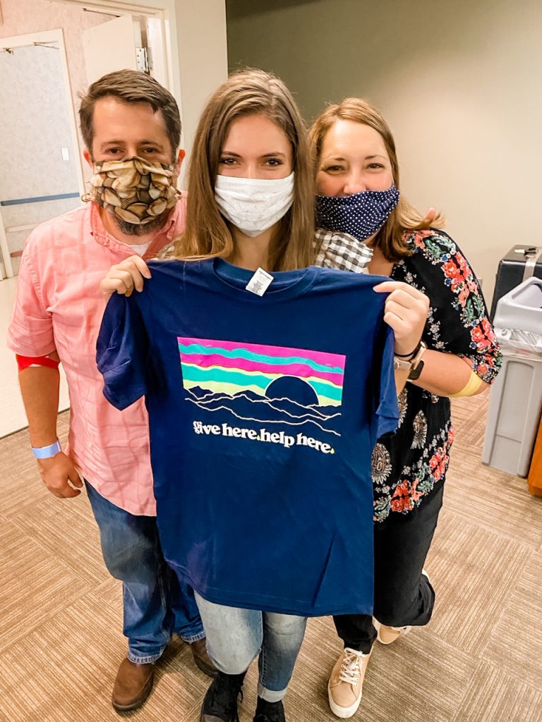 Three people wearing masks hold a T-shirt that says "Give Here, Help Here"
